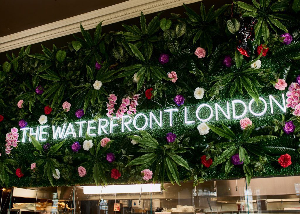 a wall logo says "the waterfront london" amidst a wall of flowers and greenery