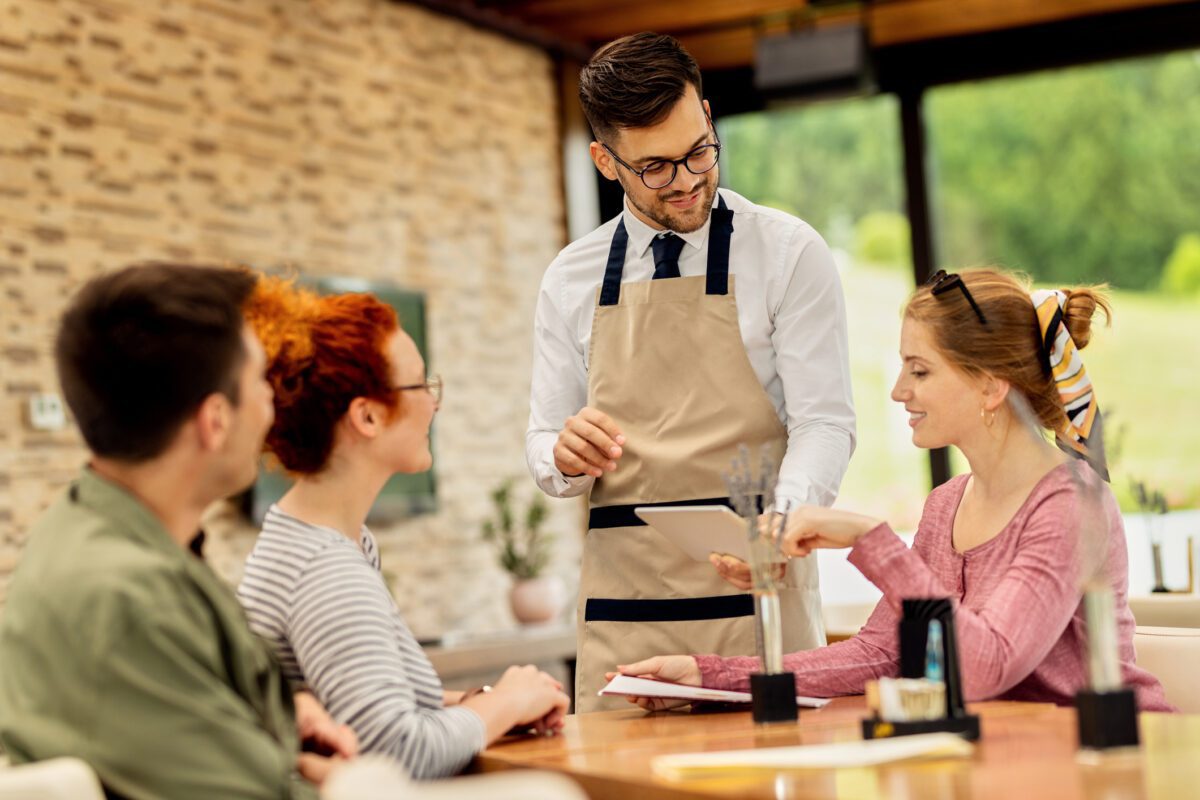 Young waiter showing menu on touchpad to his customers in a cafe.